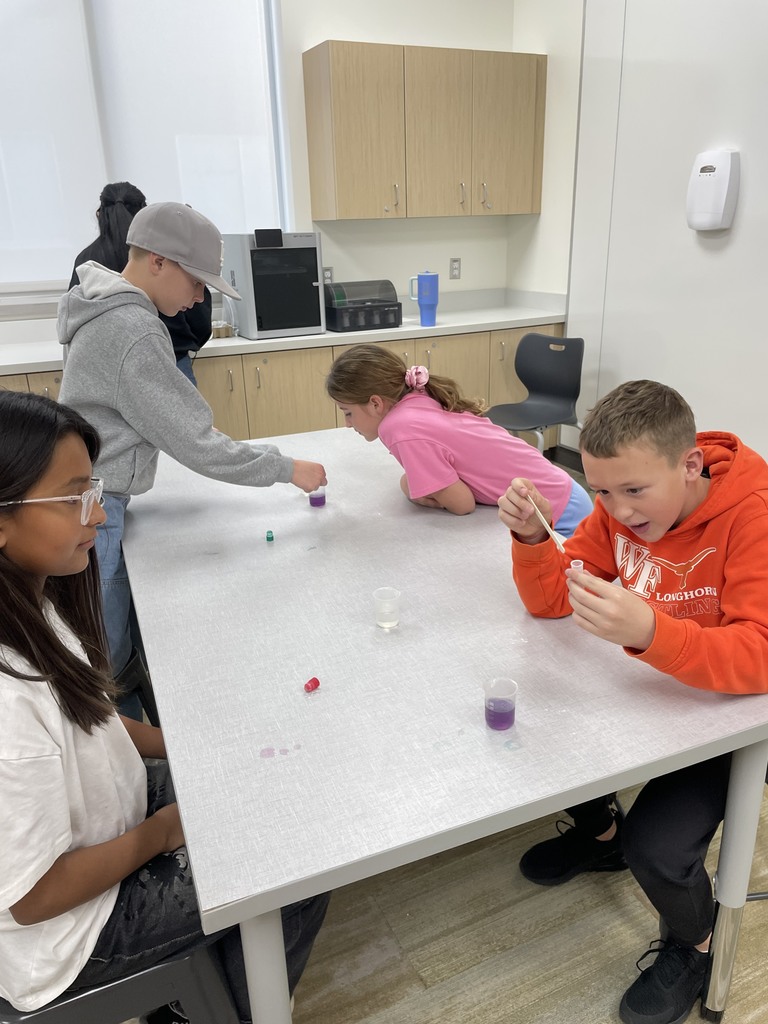 Four students, two boys and two girls, participating in a chemistry experiment in a classroom. The boy on the right, wearing an orange longhorn shirt, is looking excited while using a dropper to add a liquid to a small vial of purple liquid. Other students are leaning over the table, examining their own reactions with small vials.