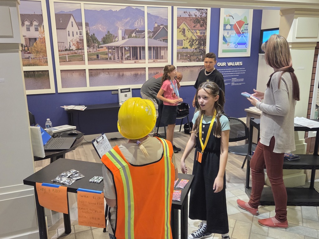 A girl wearing a construction hard hat and an orange safety vest is standing behind a small table, talking to other students. The table has signs listing prices for items, one partially visible sign reads "T-shirts $2.00...". The background wall features large, framed photographs of suburban homes and a poster about "OUR VALUES".
