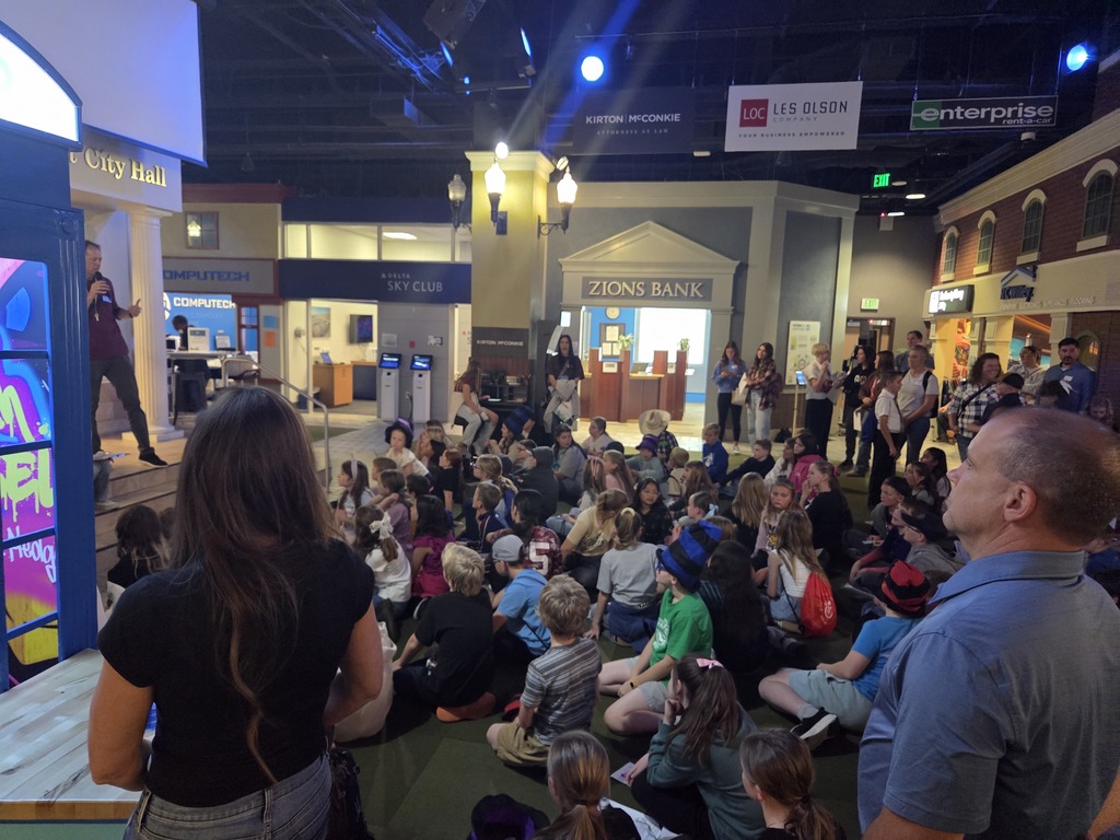 An indoor view of a simulated city environment with a large group of children and adults seated on the floor. Booths resembling storefronts like "ZIONS BANK", "DELTA SKY CLUB", and signage for "KIRTON MCCONKIE" and "LOC LES OLSON ENTERPRISE" are visible in the background. A man stands on a raised platform addressing the crowd on the left.