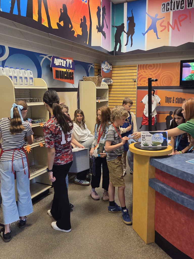 A group of students are role-playing in a simulated retail store environment. The store walls are decorated with silhouettes of people engaged in activities, and the sign on the shelving unit reads "AISLEY'S MUNCHIES". A student at the counter on the right has a sign that reads "Show Green Card HERE for 50%".