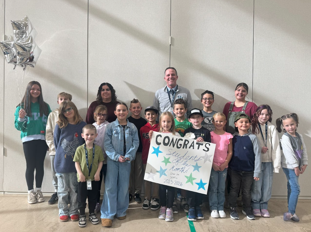 A group of 16 students, ranging in age from elementary to middle school, and two adults (one male, one female) are posing together indoors in front of a neutral-colored wall. The students are the "Students of the Month" for November 2025-2026. A man in the center, wearing a collared shirt and tie, and a woman to his left, are smiling. Several students hold a large sign that reads "CONGRATS Students Of The Month! SCAM 2025-2026" with teal and blue star graphics. A girl on the far left holds a bouquet of silver star-shaped helium balloons. The students are casually dressed and smiling.