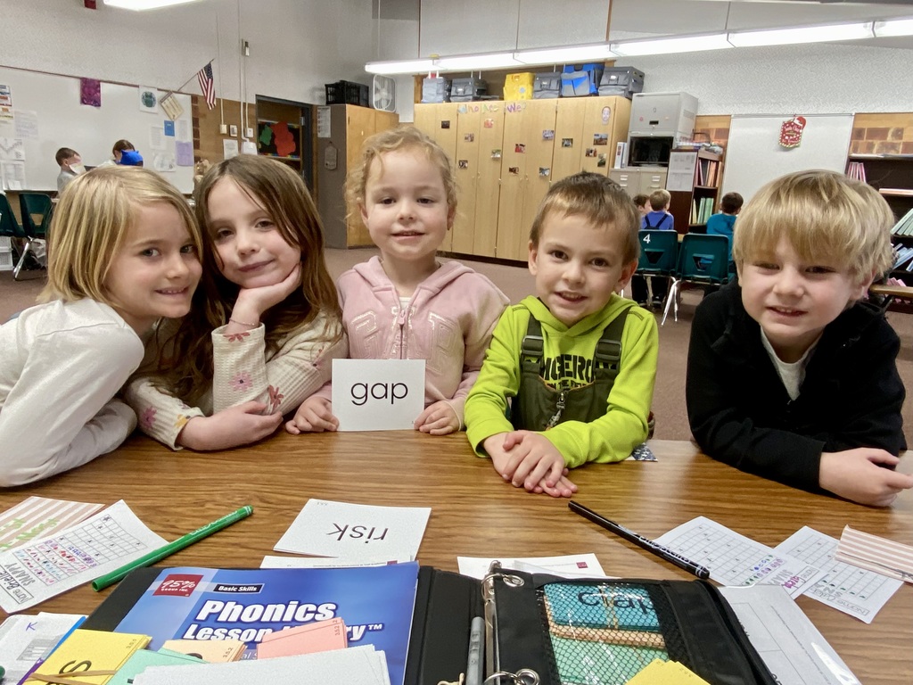 Five young elementary school students sitting together at a table, smiling at the camera. The student in the middle is holding up a flashcard with the word "gap." Various worksheets, pens, and phonics lesson materials are on the table in front of them.