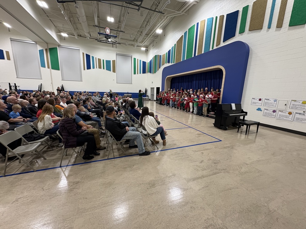 A very wide shot showing the full scale of a holiday concert in a large, brightly lit school gymnasium. A children's choir is performing on a stage with a blue backdrop and green and white wall panels. A large audience of parents and family members is seated on folding chairs spanning across the left side of the room, watching the performers. A black digital piano is visible on the right side of the stage.