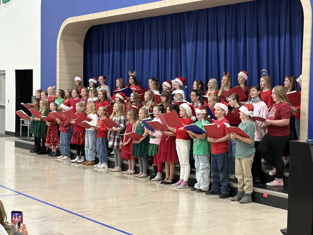 A close-up view of a large children's choir performing on a low stage. The children are dressed in various red and green holiday-themed outfits, including Santa hats and reindeer headbands, and are holding red or green songbooks. They are standing in three rows with expressions of concentration and joy.