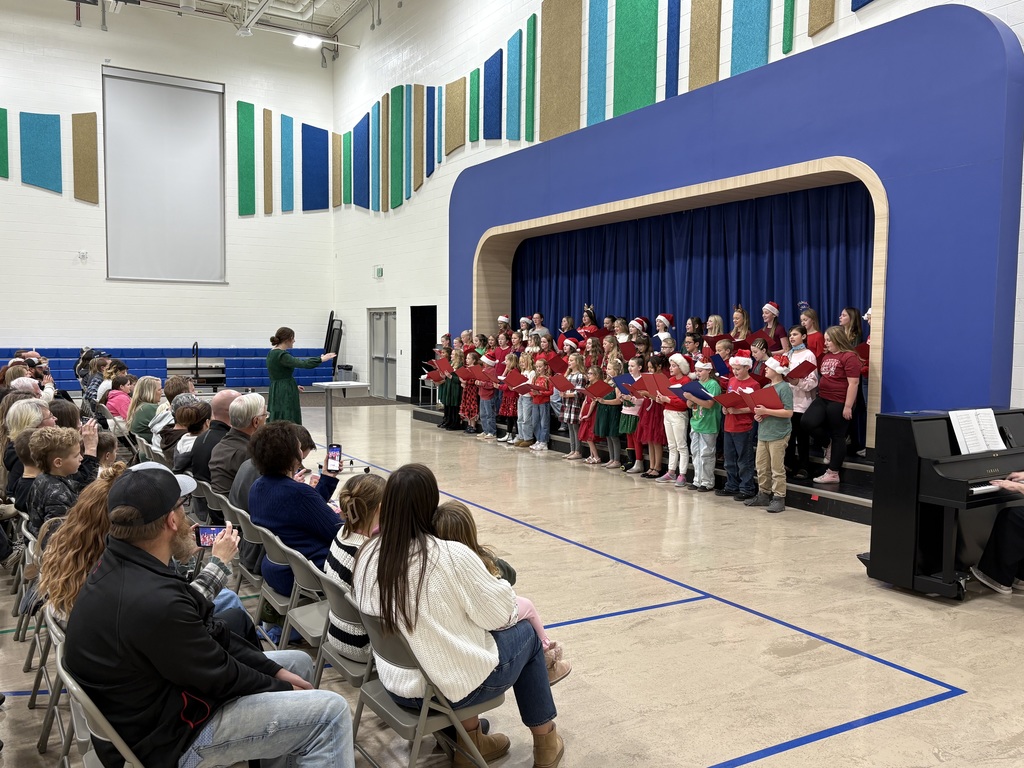 A wide-angle photo of a children's choir performing a holiday concert on a low stage in a school gymnasium. The children, dressed in red and green and wearing Santa hats and reindeer antlers, stand in three rows holding red songbooks. An adult conductor in a green dress stands to the left of the stage. A large audience of parents and families is seated on folding chairs in the foreground, watching the performance.