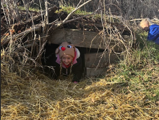 A young girl wearing a brown plush reindeer hat with a red nose smiles as she crawls out of a small, wooden underground tunnel or "burrow" covered in dry grass and straw.
