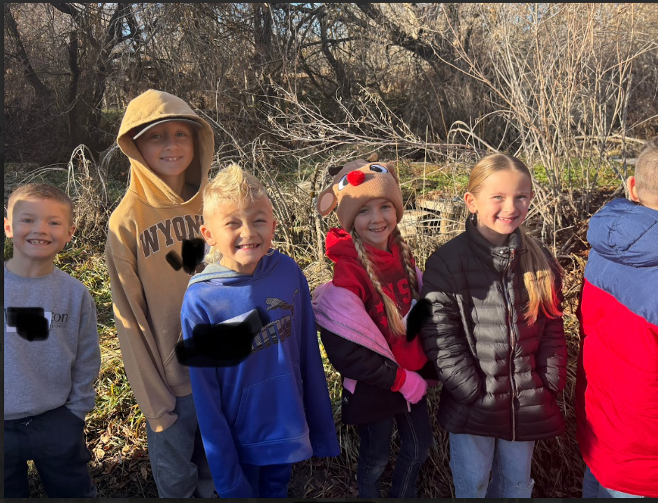 A group of five children standing outdoors in front of tall, dry brush. A girl in the center wears a plush reindeer hat and a red sweatshirt. To her left, a boy wears a tan hoodie with "WYOMING" printed across the chest.