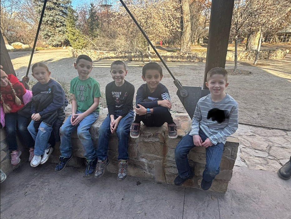 Five young boys sitting in a row on a low stone wall outdoors. The boy in the middle is wearing a black "Spider-Man" shirt, and the boy to his left wears a green shirt that says "Time to Shine." Bare trees and a circular stone walkway are visible in the background.