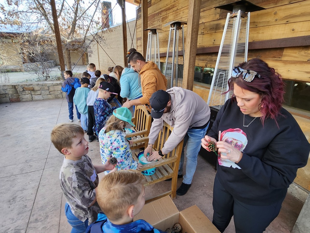 Adults and children gathered around wooden benches on a patio, working on a craft project. A woman in a black sweatshirt with a Santa graphic is inspecting a pinecone. Blue buckets containing craft supplies sit on the benches near several tall silver patio heaters.