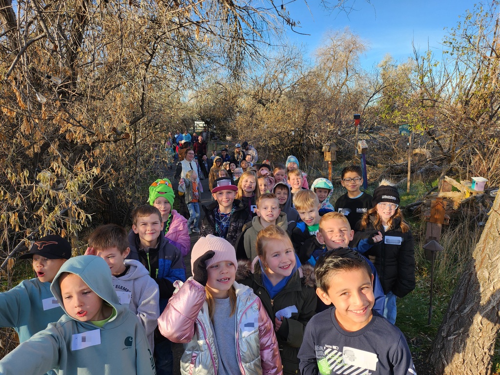 A long line of smiling children walking toward the camera on a dirt path through a wooded area. The children are wearing winter coats and hats. One boy in the foreground wears a navy long-sleeve shirt with a graphic of a battery charging. Small birdhouses are visible on posts along the trail.