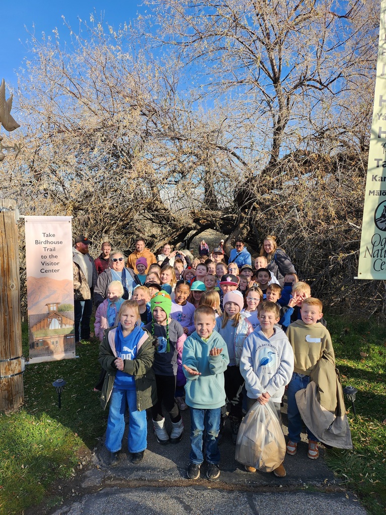 A large group of elementary school students and adult chaperones posing for a photo at the start of a trail under a natural archway of bare trees. A vertical banner on the left reads, "Take Birdhouse Trail to the Visitor Center" with an illustration of a building. A banner on the right partially shows the "Ogden Nature Center" logo.