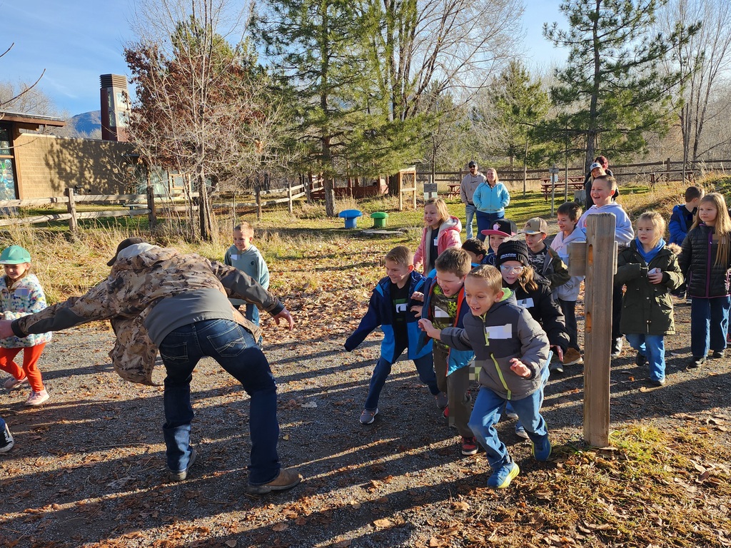 A group of children excitedly running along a gravel path toward a man in a camouflage jacket who has his arms outstretched as if to catch them. Pine trees and a wooden fence line the background under a clear blue sky.
