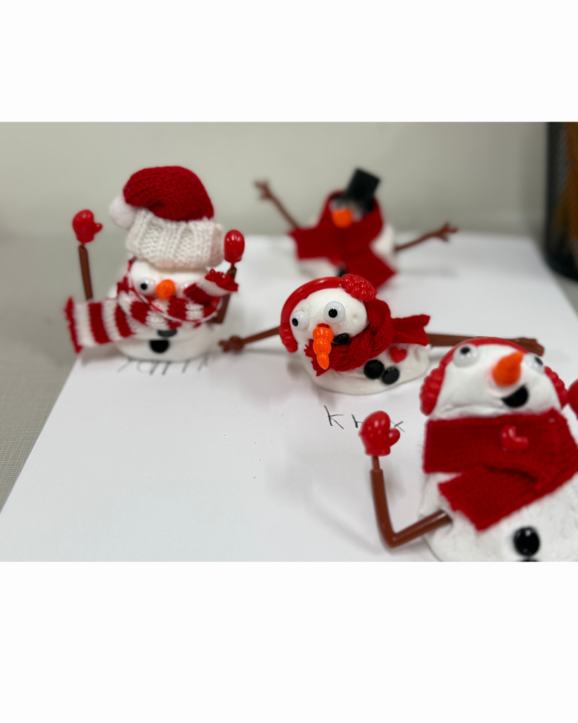 group of four unique, handcrafted clay snowmen displayed on a white surface, each wearing different red accessories like hats, scarves, and earmuffs.