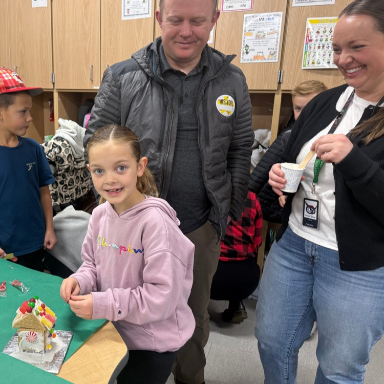 A young girl in a pink hoodie smiles while building a gingerbread house with the help of two adults in a classroom setting.