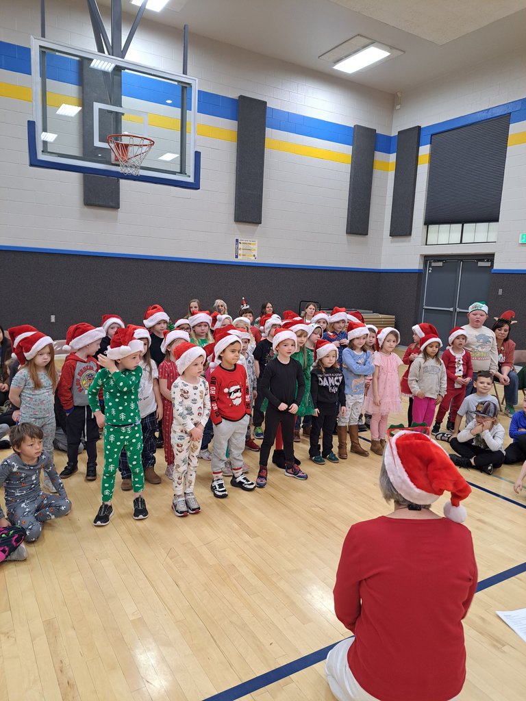 A large group of younger students standing on the gym floor in their pajamas and Santa hats, singing together.