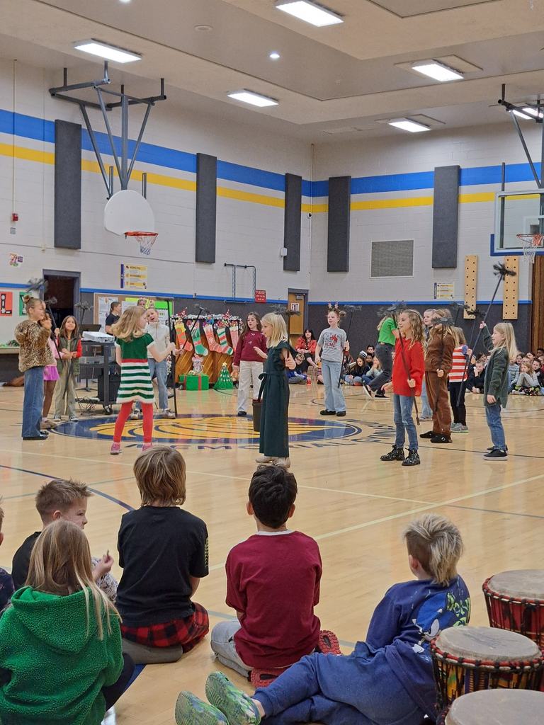 A large group of younger students standing on the gym floor in their pajamas and Santa hats, singing together.