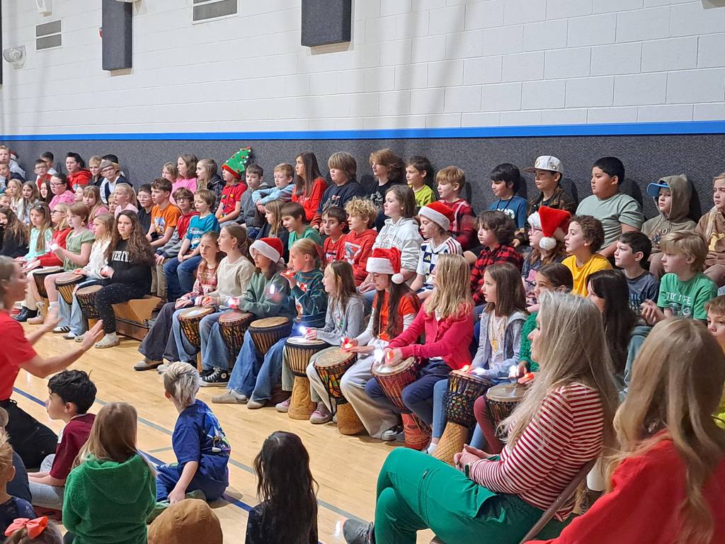 A wide shot of the gym bleachers filled with students holding djembe drums and wearing glowing finger lights.