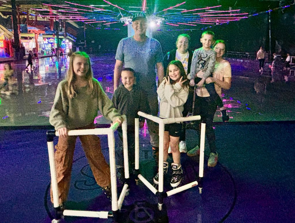A large group of children and adults posing for a photo on a roller rink floor under glowing neon lights. Two girls in the front hold white PVC skating trainers. The man in the center, Mr. Skeen, wears a shirt that says "HAVEN BAY ELEMENTARY".