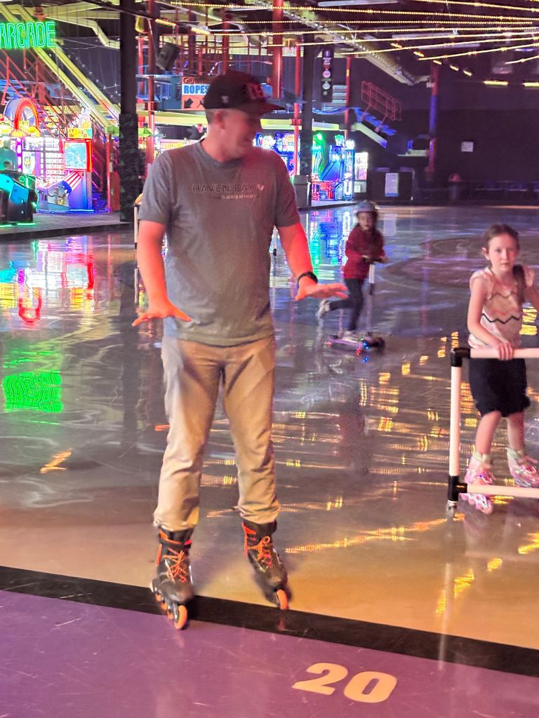 A man in inline skates, a grey shirt, and a black hat balances on a roller rink floor. Behind him, a young girl uses a skating aid. Neon arcade signs are visible in the background, including one that says "ARCADE" and another for "GRAVITY ROPES". The man's shirt says "HAVEN BAY ELEMENTARY" and the number "20" is printed on the purple floor.