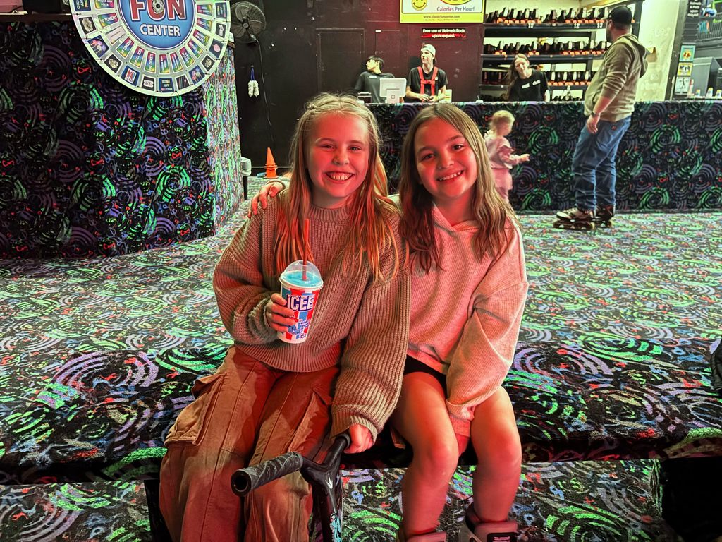Two young girls sit together on a bench at a roller rink. One girl holds a blue frozen drink in a cup labeled "ICEE". Behind them is a "Classic Fun Center" prize wheel and a counter. Signs in the background include "Calories Per Hour!" and "Free use of Helmets Pads upon request."