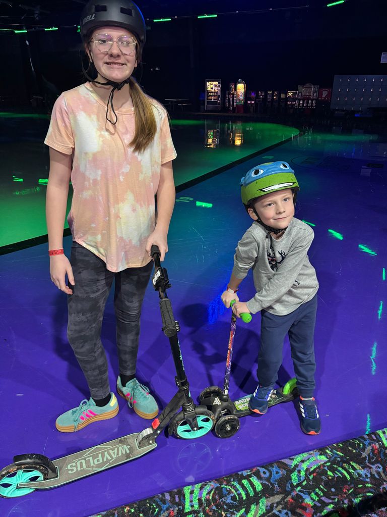 A teenage girl and a young boy pose with their scooters on a purple rink floor. The girl wears a black helmet and a tie-dye shirt, while the boy wears a Teenage Mutant Ninja Turtles helmet. The girl's scooter has the word "WAYPLUS" on the deck.