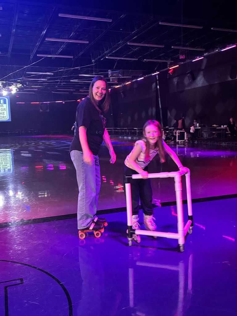 A woman in a black shirt and a young girl in a patterned shirt smile while roller skating on a rink floor lit with purple and pink lights. The girl is holding onto a white PVC skating trainer for balance. A sign in the distance contains the words "FOR THE FLOOR!".