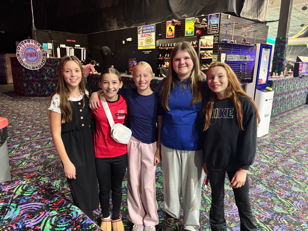 Five young girls stand in a row smiling for a group photo at a fun center. They are standing on a black carpet with a neon swirl pattern. In the background, signs read "RENT ME!", "SKATING 450", "PREMIUM", and "ROLLER BLADE RENTAL". One girl’s red shirt says "SWAT CRUSH SOCCER" and another wears a black sweatshirt with "NIKE" in rainbow letters.