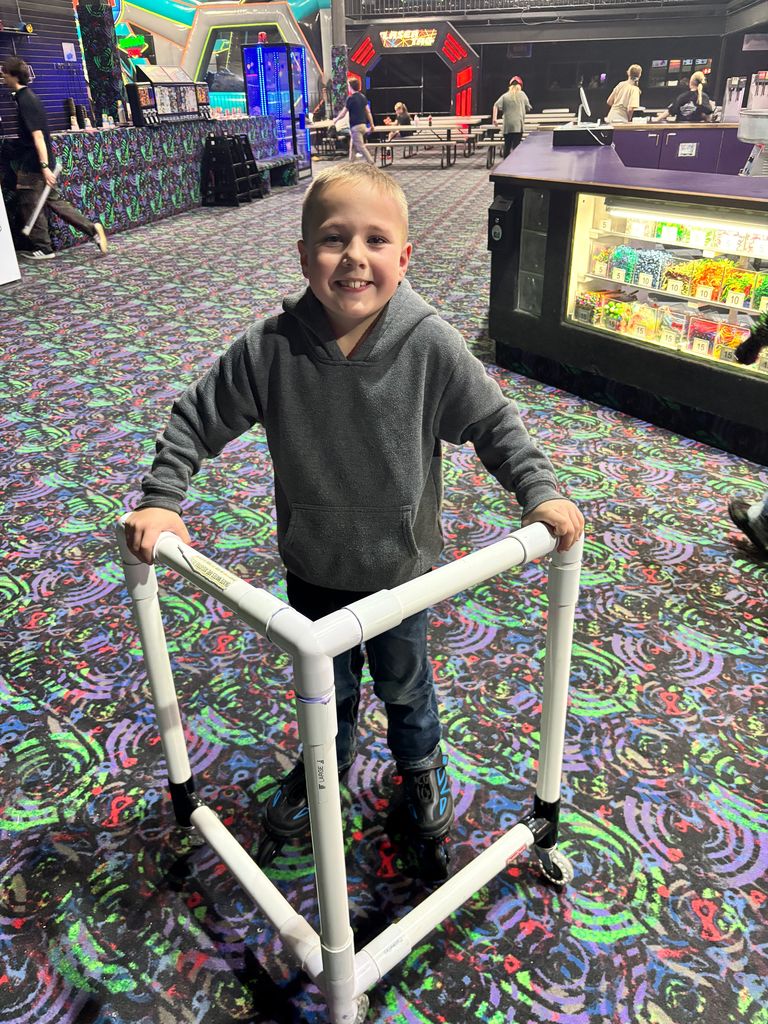 A young boy in a grey hoodie and jeans smiles brightly while using a white PVC pipe skating aid on a patterned carpet at a roller rink. In the background, there is a candy counter and a "Laser Tag" entrance. The text on the skating aid reads "SKATE MATES ARE AS-IS" and "LARGE".