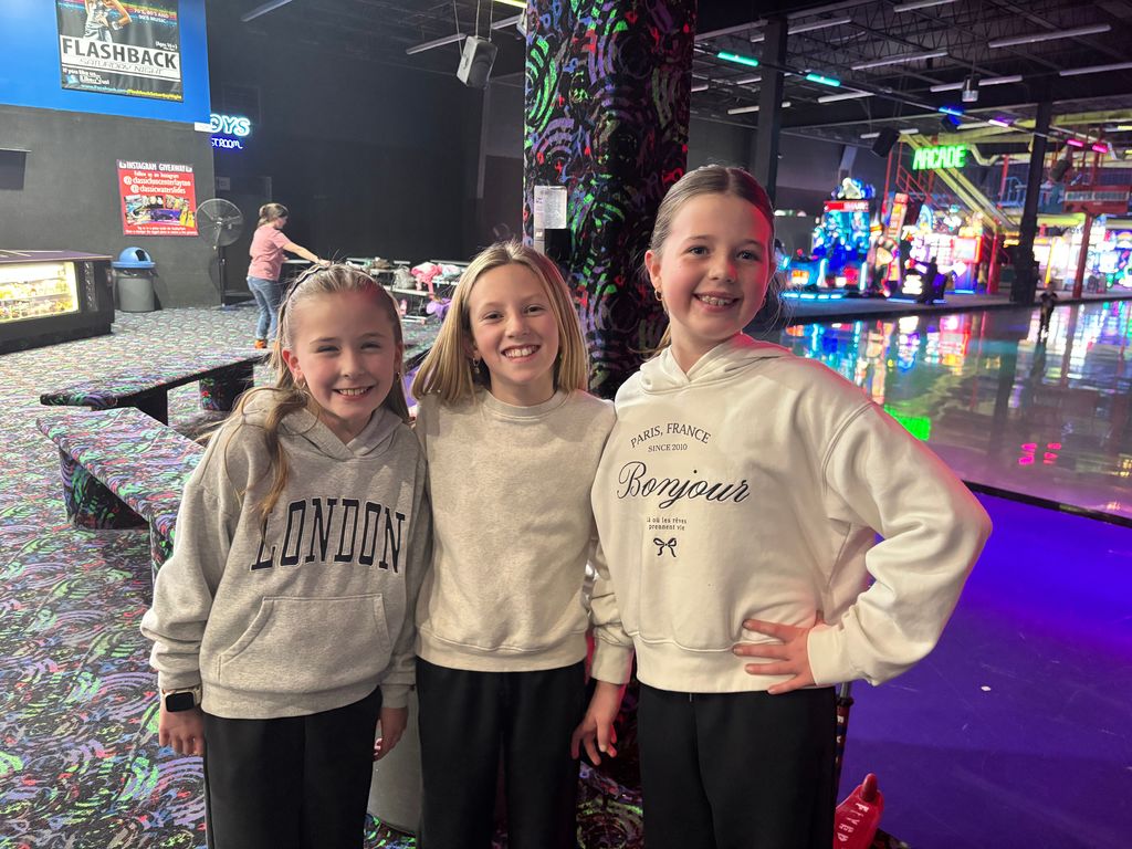 Three young girls stand together smiling in front of a colorful carpeted pillar. The girl on the right wears a white hoodie that says "PARIS, FRANCE SINCE 2010 Bonjour". The girl on the left wears a grey hoodie with the word "LONDON". A sign in the background says "FLASHBACK SATURDAY NIGHT" and "ARCADE".