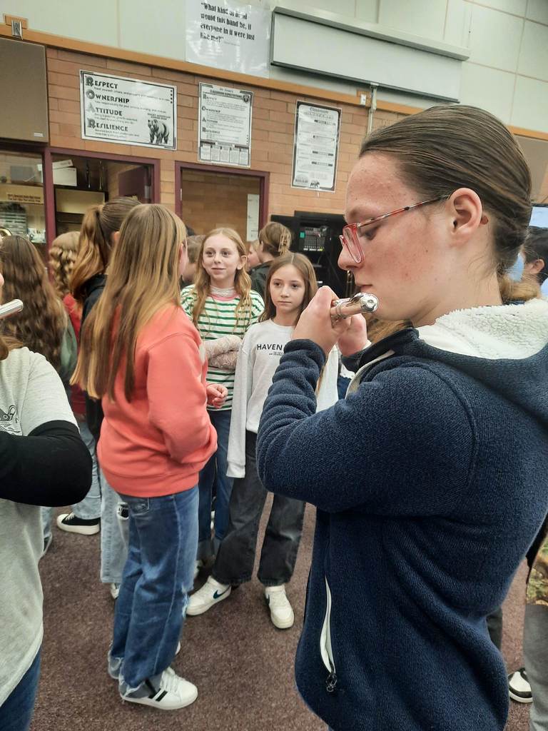 A profile view of a girl with glasses playing a flute in a crowded room. Behind her, a poster on the brick wall reads, "What kind of band would this band be, if everyone in it were just like me?" Another poster lists "RESPECT, OWNERSHIP, ATTITUDE, RESILIENCE." A student in the background wears a sweatshirt with the word "HAVEN" partially visible.