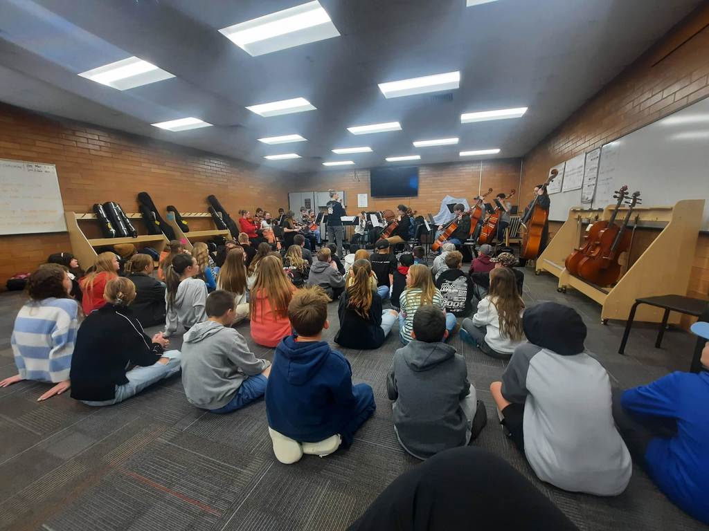 A wide shot of a school music classroom with brick walls. A large group of students sits on the floor in the foreground, watching a smaller group of students seated in chairs playing cellos and violins. A teacher stands in the center of the room. There are whiteboards on the walls and instrument storage racks along the left side.
