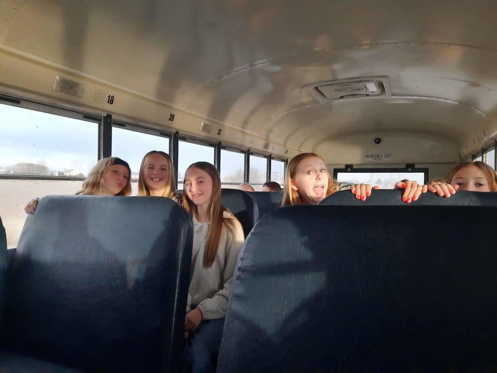 Four young girls sit together on a school bus seat, peering over the top of the blue seat back in front of them. They are smiling, and one girl is playfully sticking her tongue out. The numbers 18, 20, and 22 are printed above the windows, and the words "EMERGENCY EXIT" are visible above the rear door.