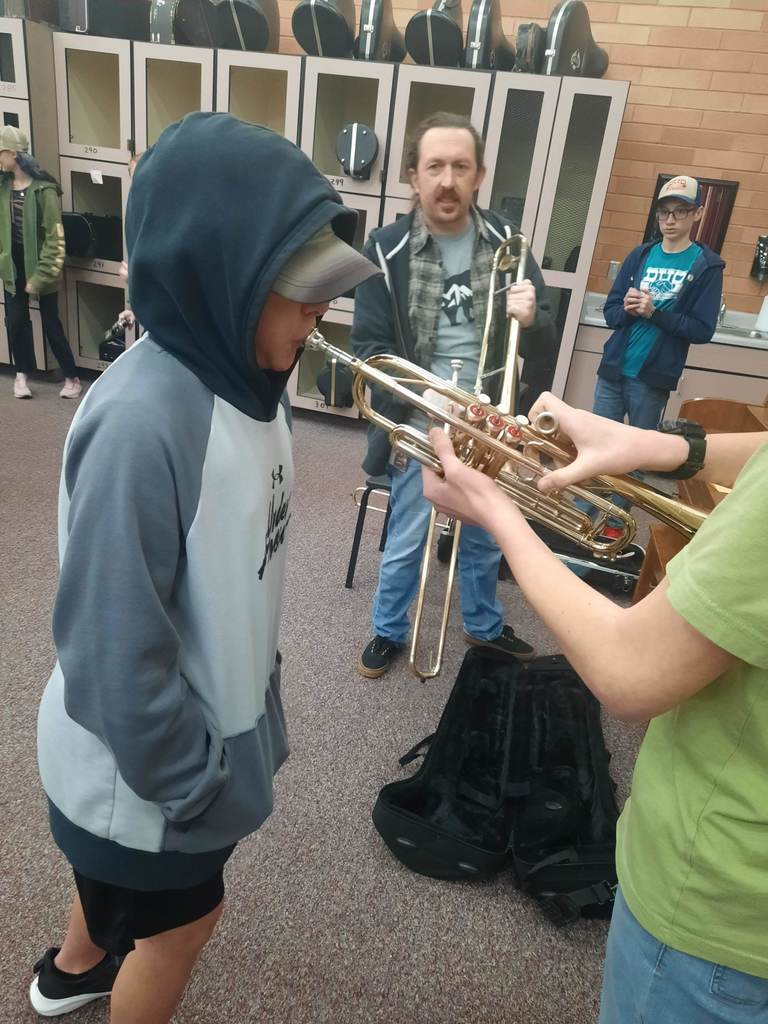 A student wearing a grey and blue hoodie with the hood up stands in a band room while someone off-camera holds a trumpet up to his mouth. An adult man holding a trombone stands behind him, and another student wearing a blue "BYU" shirt watches from the background. Beige lockers with numbers like 290 and 299 line the wall.