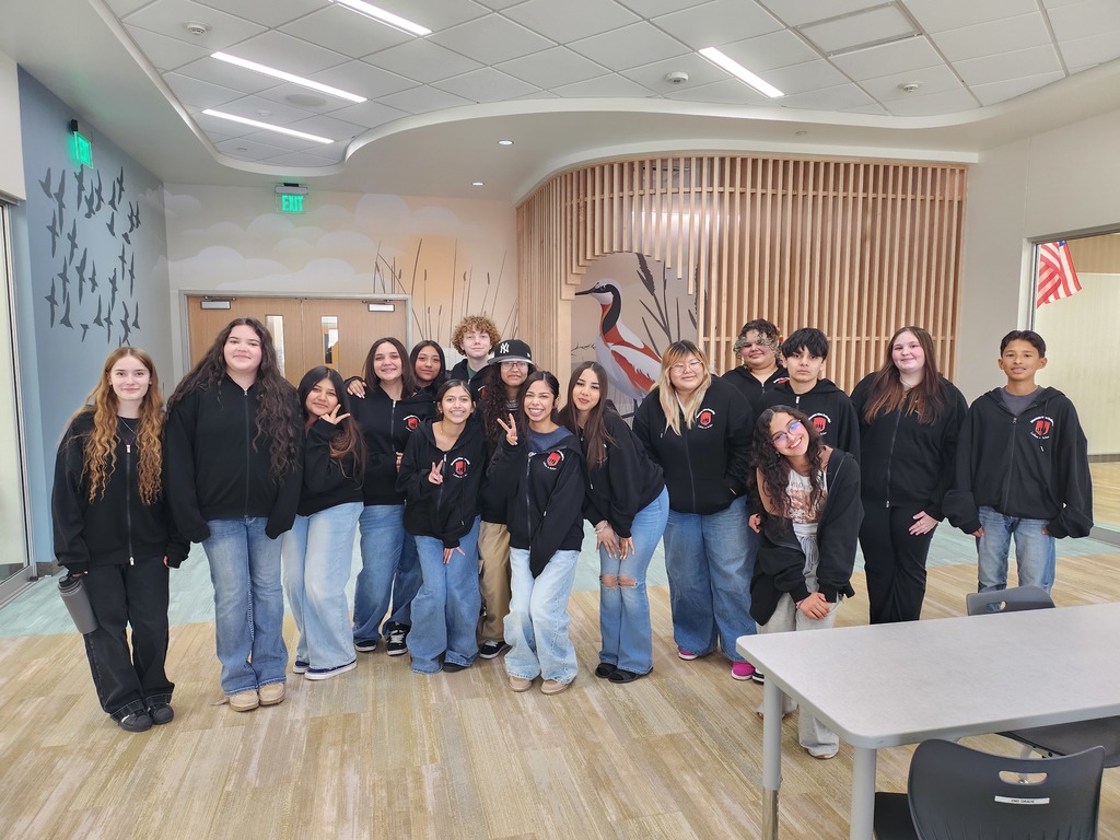 A group portrait of about fifteen teenagers standing in a modern school hallway. Most are wearing matching black zip-up hoodies with the red "Mountain West Junior High - Latinos In Action" logo. The hallway features a curved wooden slat wall and a mural of birds in flight.
