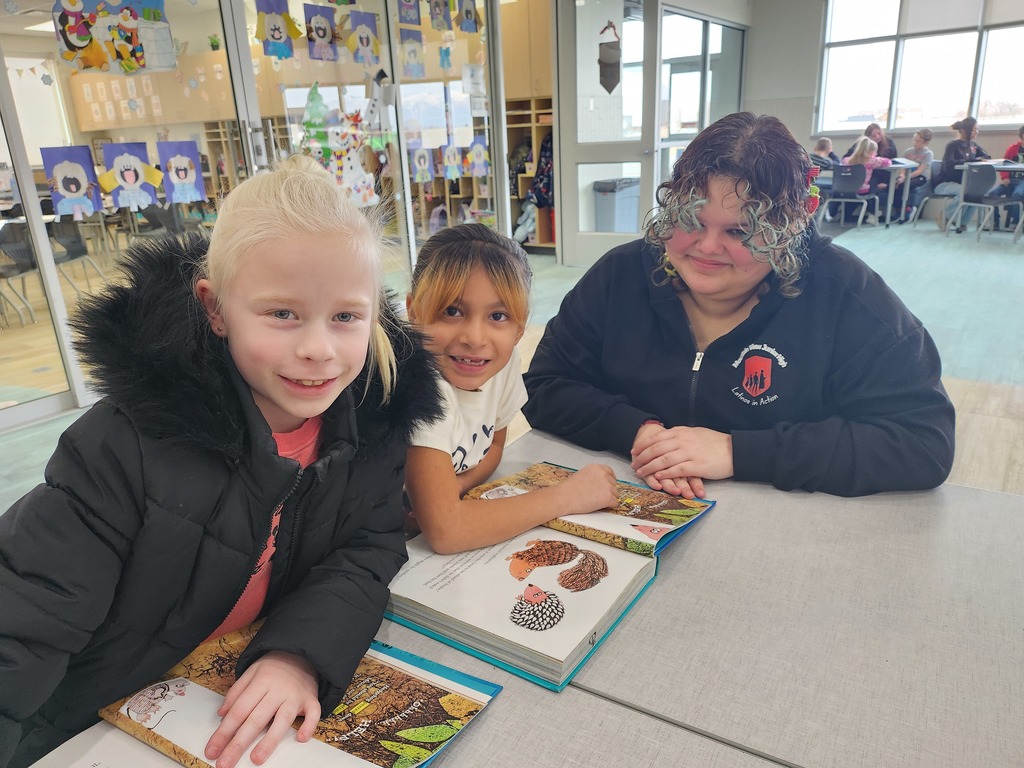 Two elementary-aged girls and an older student smile for a photo while sharing picture books at a desk. The older student wears a black zip-up hoodie with the "Mountain West Junior High - Latinos In Action" logo. The classroom background features glass windows decorated with student winter art projects.
