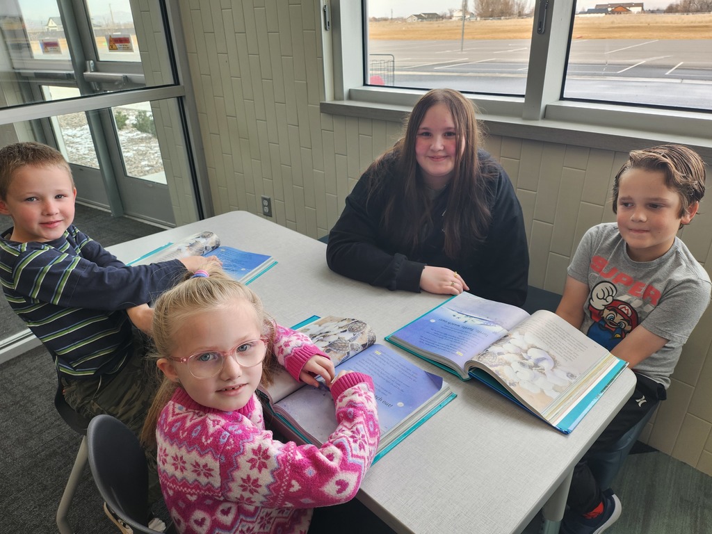 A group of four students sit at a light gray table by a large window overlooking a field. Three younger children and one older student are reading books together. One boy wears a grey shirt with a "SUPER MARIO" graphic.