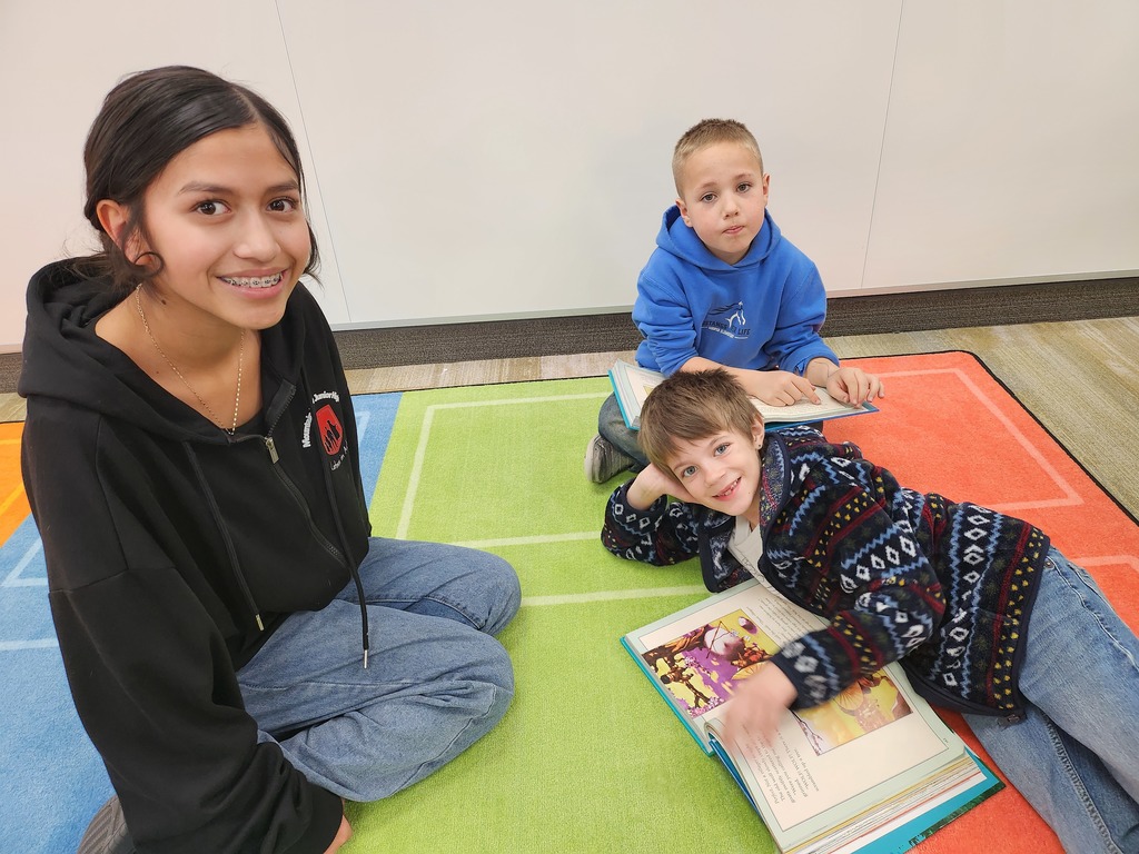 A teenage girl with braces and a black "Latinos In Action" hoodie sits on a green and blue patterned rug with two younger boys. They are reading an illustrated book together. The younger boy in the foreground is leaning on his elbow and smiling at the camera.