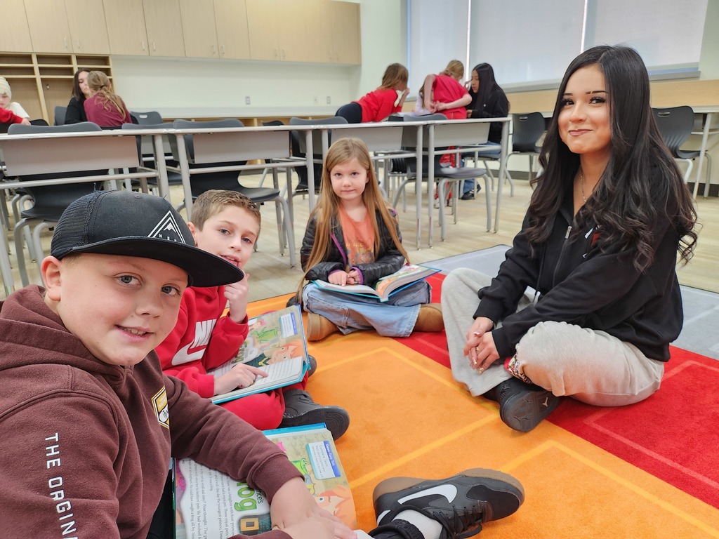 Three children and a teenage girl sit cross-legged on a bright orange and red rug in a classroom, looking at the camera. The boy in the foreground wears a brown hoodie with "THE ORIGINAL" printed on the sleeve. A picture book is open on the rug showing a story titled "The Frog."