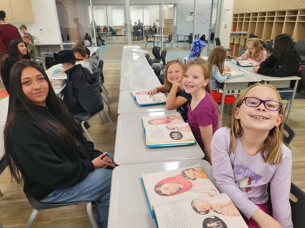 A long row of desks in a classroom where several students are seated. In the foreground, a young girl in a purple "UNICORN" shirt and glasses smiles brightly. Beside her, a teenage girl with long dark hair sits patiently. Multiple copies of a picture book with an illustration of a woman in a red headscarf are open on the desks.