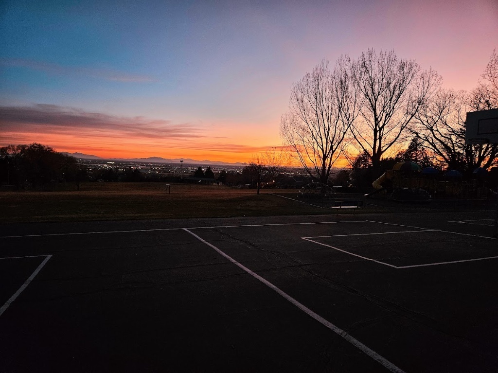 A wide-angle view of a basketball court and playground at Lomond View Elementary during a vibrant sunset. The sky transitions from deep orange and gold at the horizon to a soft purple and blue above. In the background, the city lights and mountain silhouettes are visible under the glowing sky.
