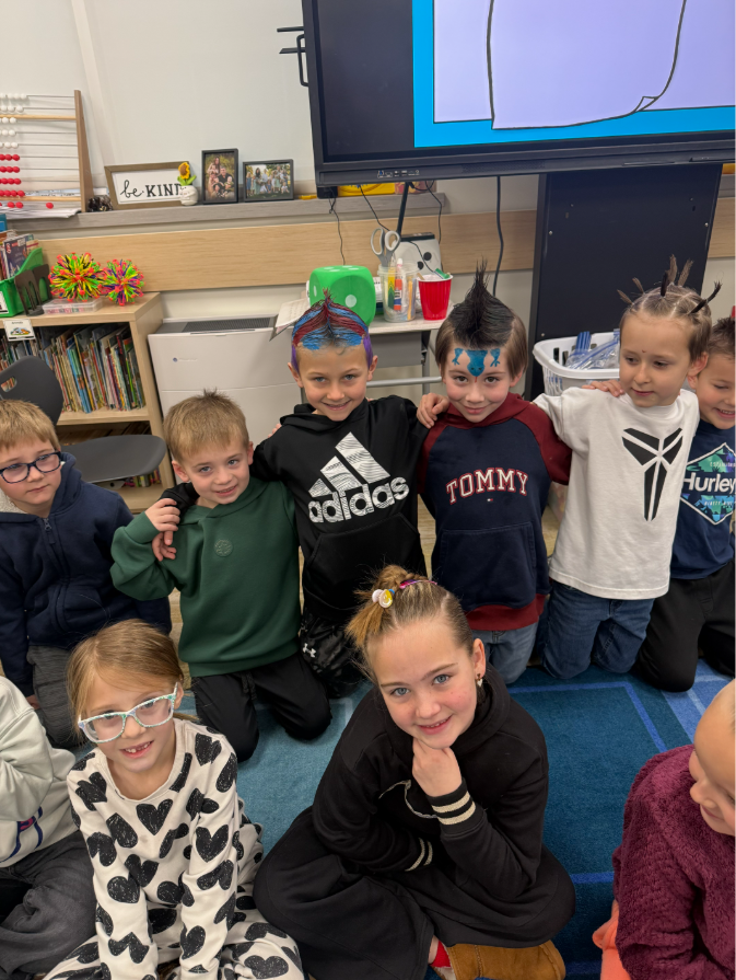 A group of students kneeling on a blue rug. The boy in the center has hair dyed blue, red, and purple in a mohawk style and wears an "adidas" hoodie. The boy next to him has a dark mohawk and a blue lizard or frog painted on his forehead, wearing a "TOMMY" hoodie. A girl in the front wears a white shirt with a black heart pattern. A "be KIND" sign and a wooden abacus are visible on the shelf behind them.