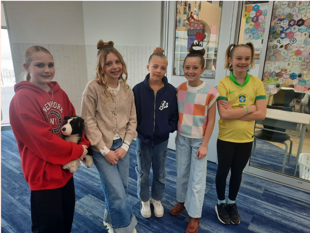 Five young girls standing in a school hallway showcasing creative "Crazy Hair Day" styles. One girl on the far left wears a red hoodie with "NEW YORK CITY" and "NY" printed on it, holding a stuffed border collie. The other girls feature hairstyles including a hair-bow made of hair, a "poop emoji" bun with googly eyes and a red heart, and high pigtails. A girl on the right wears a yellow Brazil soccer jersey with the CBF Brasil crest. 