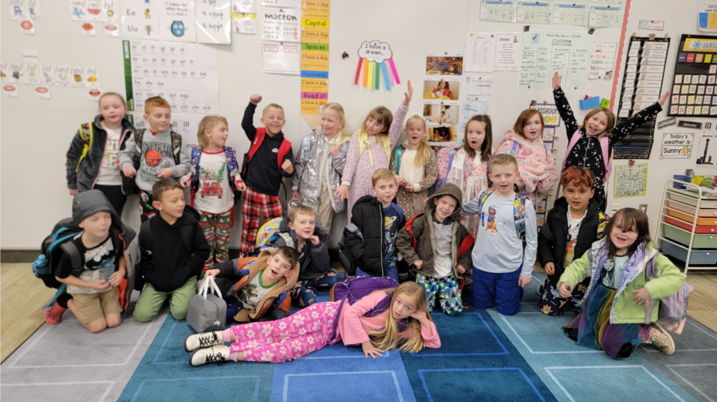 A group of about 20 students in pajamas sitting and standing on a blue patterned rug in a classroom. One girl in the foreground lies on her side wearing pink "Barbie" silhouette pajama pants. The room is decorated with educational posters, including a "Today's Weather is Sunny" sign, a "I have a dream..." rainbow decoration, and a "Classroom Expectations" chart.
