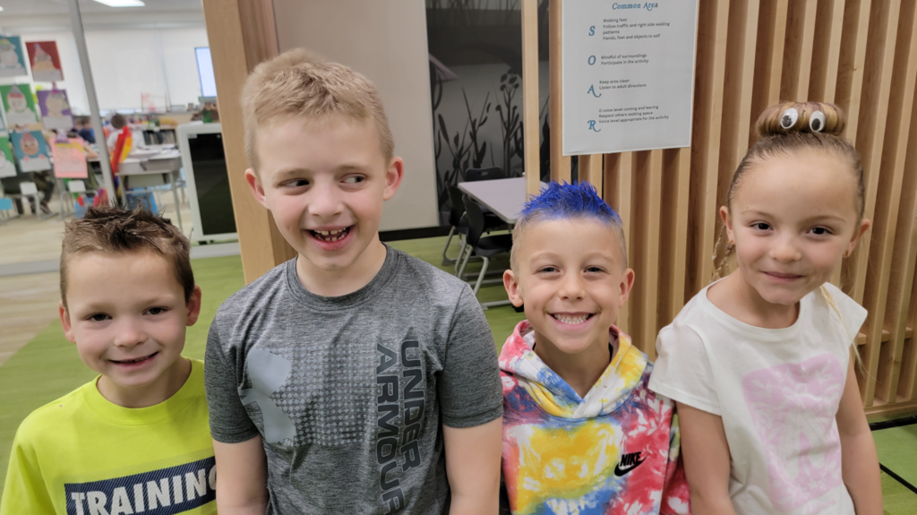 Four elementary students posing in a school common area. The boy in the center-right has bright blue dyed hair and wears a colorful tie-dye Nike hoodie. The girl on the far right has her hair in a bun styled to look like a face with large googly eyes. Behind them, a "Common Area" sign lists "SOAR" expectations: "S - Safe, O - Ownership, A - Achiever, R - Respectful."