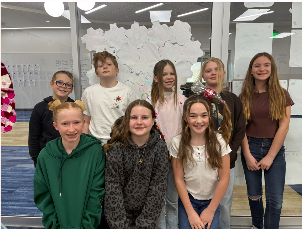 Eight older elementary or middle school students posing in front of a glass wall decorated with hexagonal paper cutouts, one of which has the name "CASH" written on it. Creative hairstyles include a girl with a "Dr. Pepper" soda bottle incorporated into a high ponytail to look like the liquid is pouring out. Other students wear hoodies in green, grey, and black, including one girl in a grey leopard print hoodie.