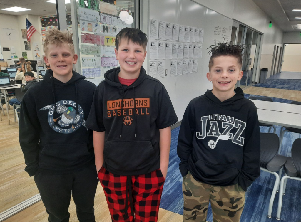 Three boys standing together in a modern classroom setting. The boy on the left wears a black hoodie with a duck logo and the text "GHG DECOY SYSTEMS." The middle boy wears a black short-sleeve hoodie over a red shirt with "LONGHORNS BASEBALL" and an "H" logo, paired with red and black buffalo plaid pajama pants. The boy on the right has spiked hair and wears a black "UTAH JAZZ" basketball hoodie and camouflage pants.