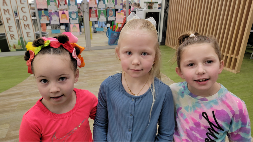 Close-up portrait of three young girls in a classroom. The girl on the left has her hair styled in multiple small buns adorned with several bright pink, orange, yellow, and green bows; she wears a bright pink Nike shirt. The middle girl has blonde hair with a white lace bow. The girl on the right has a high ponytail and wears a tie-dye shirt with the word "JUST" visible. In the background, a sign reads "WELCOME" and "CAMPERS."
