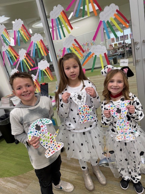 Three children stand in front of a glass wall decorated with rainbow-colored "I have a dream" crafts. They are holding paper Dalmatian cutouts covered in multi-colored spots. The girls are wearing white outfits with black spots and tutus, and one wears a spotted headband.