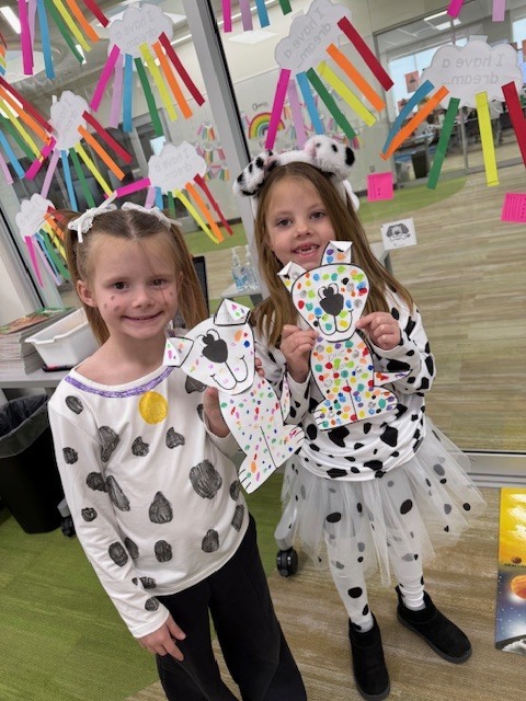 Two young girls smile while holding up their decorated paper Dalmatian crafts. Both are dressed in Dalmatian-themed clothing; the girl on the right wears a spotted headband and a white tutu. The glass wall behind them is covered in paper cloud crafts that say, "I have a dream..."