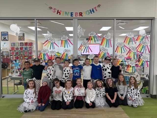 A large group of elementary students poses together in front of a glass classroom wall. Many children are wearing Dalmatian-themed costumes, spotted shirts, and face paint. A student in the center holds a sign that says, "101st Day of School." A banner above the glass reads, "SHINE BRIGHT," and the glass is covered in numerous crafts labeled, "I have a dream..."